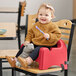 A baby sitting in a red Carlisle booster seat at a table holding a spoon.