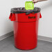 A person's hand throwing a green square object into a red Continental 44 gallon round trash can.
