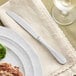 An Acopa Brigitte stainless steel dinner knife on a napkin next to a plate with meat and broccoli and a glass of wine.