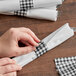 A person's hands using a black Gingham Self-Adhering Paper Napkin Band to hold folded black and white checkered napkins.