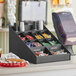 A black ServSense countertop condiment organizer with sections holding condiments on a counter.