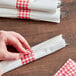 A person's hand using a red and white checkered paper napkin band to put a napkin on a table.