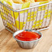 A basket of french fries with a fluted stainless steel sauce cup of red sauce on a white background.