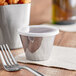 A Tablecraft stainless steel sauce cup with a white lid next to a fork on a napkin.