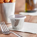A close-up of a Tablecraft stainless steel sauce cup with a plastic lid and a fork on a table.