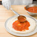 A person using a Vollrath Ivory Solid Round Spoodle to put red sauce on pizza dough.
