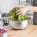 A person in gloves mixing greens in a Choice stainless steel mixing bowl with a silicone bottom.