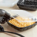 A gloved hand with a spoon putting macaroni and cheese into a black plastic Choice microwavable container.