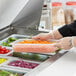 A person in gloves holding a Vigor translucent polypropylene food pan of vegetables at a salad bar.
