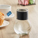 A Tablecraft glass bottle with a black cap on a table next to a cupcake and a cup of tea.