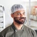 A man in a chef's uniform and beard wearing a white disposable hair net.