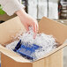 A hand putting a blue box of Spring-Fill Very Fine Paper Shred into a cardboard box filled with shredded paper.