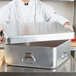A chef in a white coat holding a Vollrath aluminum double roaster pan with a lid.