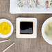 A square white porcelain sauce cup with yellow sauce on a table with sushi.