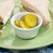 A sandwich on a plate with a white Thunder Group ramekin filled with pickles.