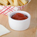 A white Tuxton china ramekin filled with french fries on a counter with a bowl of ketchup.