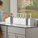A woman using a ServSense stainless steel quadruple condiment dispenser on a counter in a kitchen.