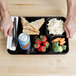 A Carlisle black 6 compartment tray with a sandwich, fruit, and a can of soda.