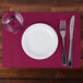 A white plate with a glass of water and a fork and knife on a burgundy Hoffmaster paper place mat with a scalloped edge.