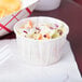 A white Solo paper souffle cup filled with coleslaw on a white counter with a bowl of potato chips.