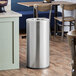 A Lancaster Table & Seating stainless steel round decorative waste receptacle on a wood floor.