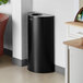 A black Lancaster Table & Seating half round decorative waste receptacle in a corner of a room.