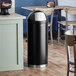 A black Lancaster Table & Seating decorative waste receptacle with a push door lid.