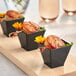 A group of small black Choice square plastic shot glasses filled with food on a table.