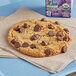 A chocolate chip cookie on a blue tray next to a bag of HERSHEY'S Semi-Sweet 1M Baking Chips.