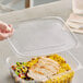 A hand holding a World Centric clear plastic lid over a deli container of food.