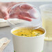 A hand holding a World Centric clear plastic container of soup with a World Centric clear plastic domed lid