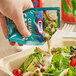 A hand pouring Kraft Creamy Caesar dressing from a packet over a salad.