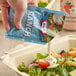 A hand holding a Kraft Ranch Dressing packet pouring dressing onto a salad.