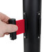 A hand holding a black Aarco crowd control stanchion with dual red retractable belts.
