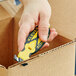 A hand using a Klever Kutter X-Change yellow and black box cutter to cut a box.