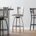 A close-up of a Lancaster Table & Seating clear coat finish swivel bar stool with a dark gray vinyl padded seat.