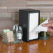 A hand holding a white napkin in a black Tablecraft Tallfold Napkin Dispenser.