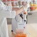 A person in a white coat and gloves uses the AvaMix food processor to shred carrots into a plastic container.