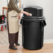 A woman wearing an apron putting a plastic bag into a black Rubbermaid trash can.