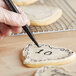 A hand using a Chefmaster black food decorating pen to draw on a heart shaped cookie.