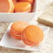 A white plate of orange macaroons with a white bowl of Chefmaster Natural Sunset Orange Liqua-Gel Food Coloring.