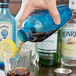 A hand using a Fineline blue plastic shaker to pour a brown drink into a glass with a lemon wedge.