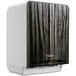 A white paper towel dispenser with a wood patterned faceplate.
