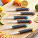 A Schraf serrated paring knife with a black TPRgrip handle cutting a lime on a wooden cutting board with a variety of colorful fruit.