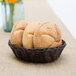 A Tablecraft brown rattan bread basket filled with bread on a table.