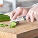 A person with plastic gloves using a Schraf smooth edge paring knife with a yellow handle to cut a cucumber on a cutting board.