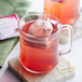 A glass mug with a Twinings Pomegranate & Raspberry Herbal Tea bag steeping in it.