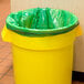 A yellow trash can with a 55 gallon eco-friendly trash bag with a green lid.