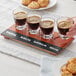 An Acopa flight tray holding four espresso glasses filled with brown liquid on a table with cookies.