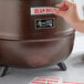 A hand pressing a label onto a copper Avantco soup kettle.